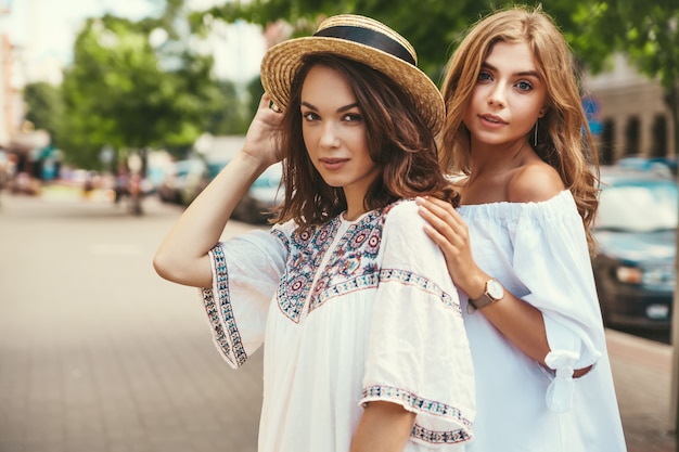 Modeporträt von zwei jungen stilvollen Hippie-Brünetten- und blonden Frauenmodellen im sonnigen Sommertag in der weißen Hipster-Kleidung, die aufwirft. Kein Make up