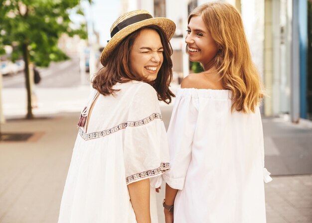 Modeporträt von zwei jungen stilvollen Hippie-Brünetten- und blonden Frauenmodellen ohne Make-up im sonnigen Sommertag in der weißen Hipster-Kleidung, die aufwirft. Dreh dich um