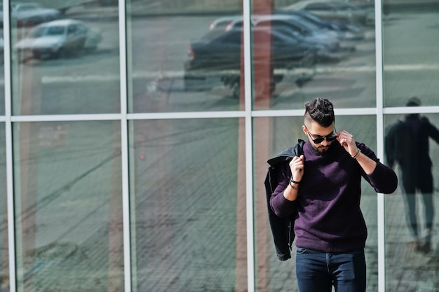 Modereicher Bart arabischer Mann trägt Rollkragen und Sonnenbrillen, die vor großen Fenstern eines modernen Gebäudes mit Jacke zur Hand stehen Stilvoller, erfolgreicher und modischer arabischer Modelltyp