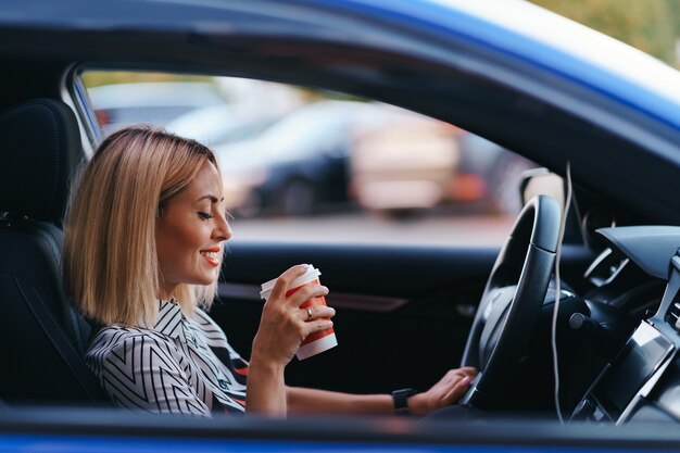 Moderne blonde Frau, die einen Kaffee zu gehen geht, während in der Stadt fährt