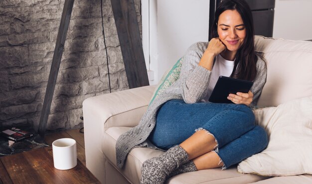 Moderne Frau, die auf dem Sofa unter Verwendung einer Tablette sitzt