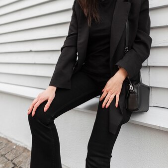 Moderne frau in modischen jeanshosen in alltäglichen stylischen ledersneakern in trendiger jacke mit schwarzer handtasche in der nähe der grauen wand in der stadt. weiblicher körper der nahaufnahme. lässige jugendmode für damen.