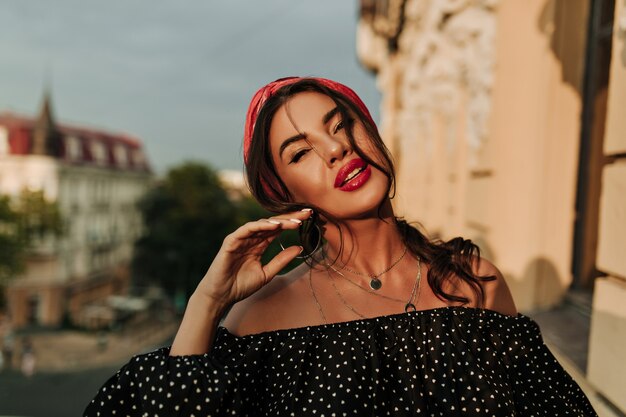 Moderne helle Dame mit schönem Make-up, rosafarbenem Stirnband und weißer Maniküre in gepunktetem schwarzem Hemd mit Blick in die Kamera auf dem Balkon..