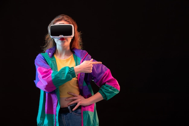 Moderne junge Dame der Vorderansicht im bunten Mantelorangen-T-Shirt, das virtuelle Realität auf dem interaktiven Spiel des schwarzen Hintergrundspiels spielend spielt