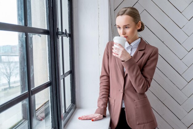 Moderne junge Frau in einer braunen Jacke, die am Fenster in einem Raum Kaffee trinkt.