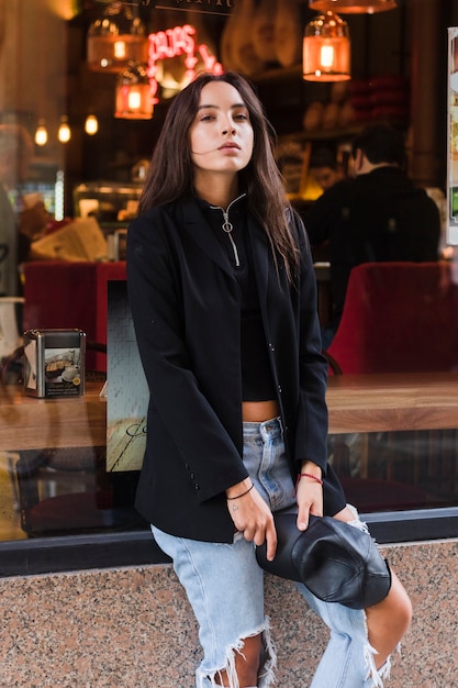 Kostenloses Foto moderne junge frau, welche die kappe in der hand sitzt vor fensterrestaurant hält