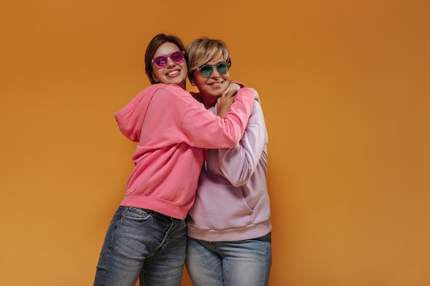 Moderne kurzhaarige Damen mit geschnittenem Lächeln und rosa und grüner Brille in den breiten stilvollen Kapuzenpullis auf orange lokalisiertem Hintergrund.