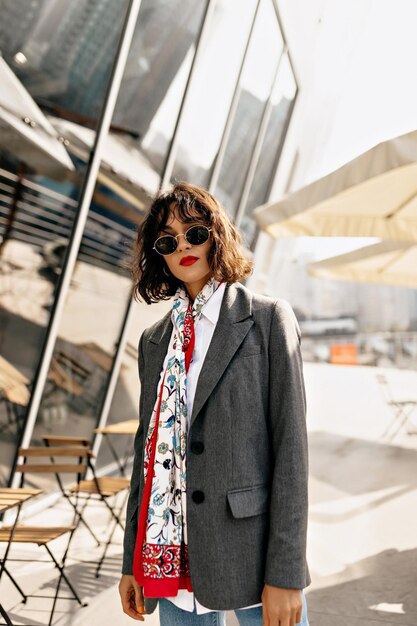 Moderne modische Frau mit Hemdfrisur in Sonnenbrille mit roten Lippen in grauer Jacke und hellem Seidenschal posiert draußen auf sonniger Straße