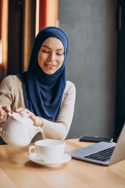 Moderne muslimische Frau, die Tee trinkt und in einem Café am Computer arbeitet