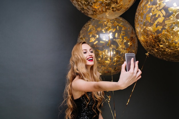 Moderne Partyzeit der jungen herrlichen Frau im schwarzen Luxuskleid, mit langen lockigen blonden Haaren, die Selfie mit großen Luftballons voll mit goldenen Lametta machen. Feiern, lächeln.