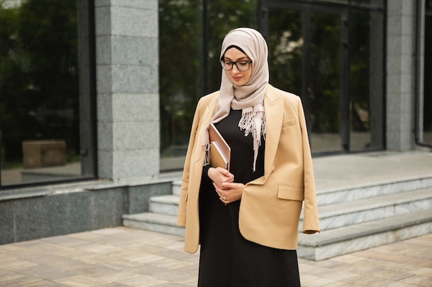 Moderne, stilvolle muslimische Frau in Hijab, Business-Stil-Jacke und schwarzer Abaya, die mit Laptop in der Stadtstraße spaziert