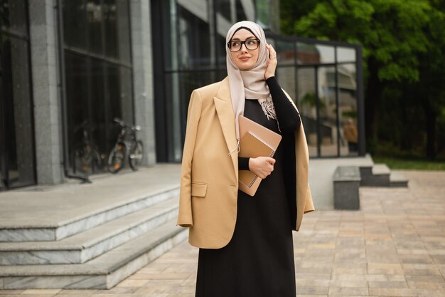 Moderne, stilvolle muslimische Frau in Hijab, Business-Stil-Jacke und schwarzer Abaya, die mit Laptop in der Stadtstraße spaziert