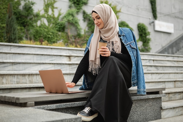 Moderne, stilvolle muslimische Frau in Hijab, Jeansjacke und schwarzer Abaya, die in der Stadtstraße sitzt und am Laptop arbeitet