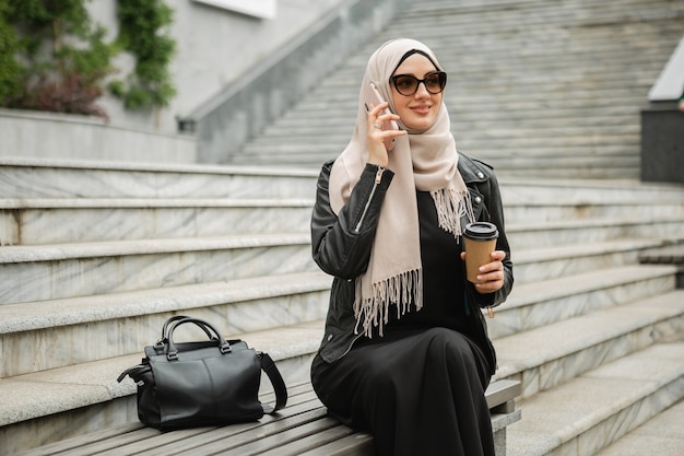 Moderne, stilvolle muslimische Frau in Hijab, Lederjacke und schwarzer Abaya, die in der Stadtstraße sitzt und mit dem Handy in Sonnenbrille spricht talking