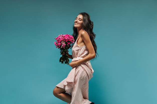Moderne süße Frau mit gewelltem Haar in kariertem, stilvollem Sommerkleid mit rosa Blumenstrauß und lächelnd auf isoliertem blauem Hintergrund