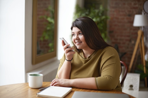 Moderne Technologie, Kommunikation und Lifestyle. Entzückende schöne junge brünette Frau mit kurvigem Körper, der Sprachnachricht über Online-Messenger-Anwendung unter Verwendung des Mobiltelefons am Kaffeetisch aufzeichnet