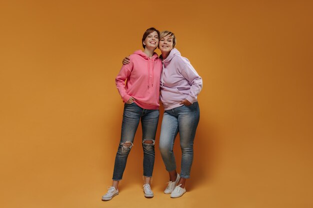 Moderne zwei Frauen mit cooler kurzer Frisur in weiten rosa Kapuzenpullis, Röhrenjeans und weißen Turnschuhen, die in die Kamera schauen und lächeln.