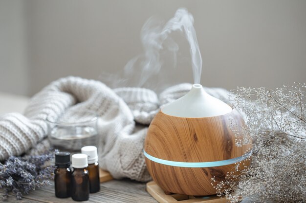 Moderner Aromaöldiffusor auf Holzoberfläche mit Strickelement, Wasser und Ölen in Gläsern.