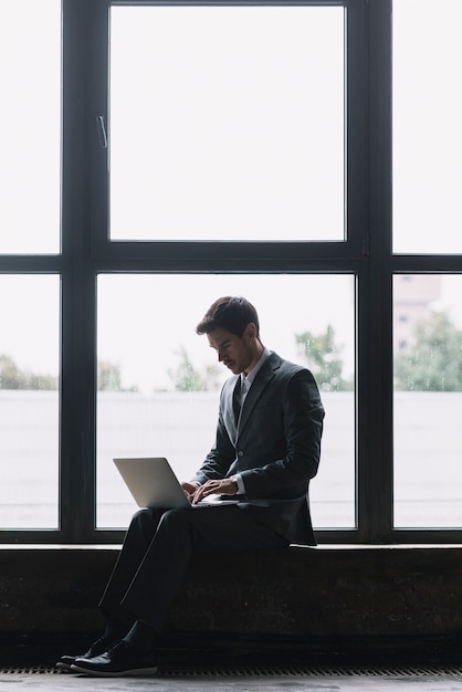Moderner Geschäftsmann mit Laptop auf seinem Schoss sitzend vor Fenster