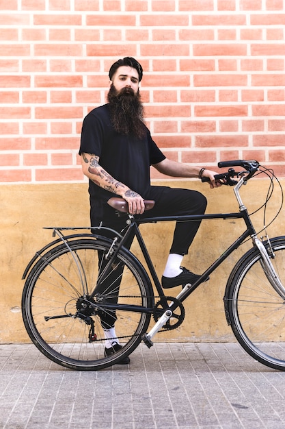 Moderner junger Mann, der mit seinem Fahrrad gegen Backsteinmauer steht