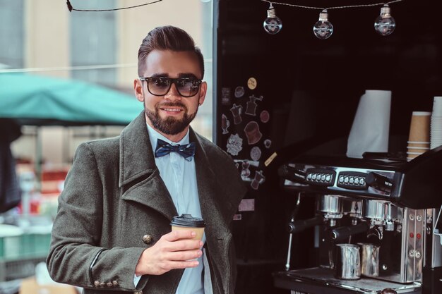 Moderner lächelnder Mann mit Sonnenbrille trinkt Kaffee, während er draußen im Coffeeshop steht.
