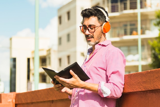 Moderner Mann, der gegen hörende Musik der Wand auf Kopfhörerlesebuch steht