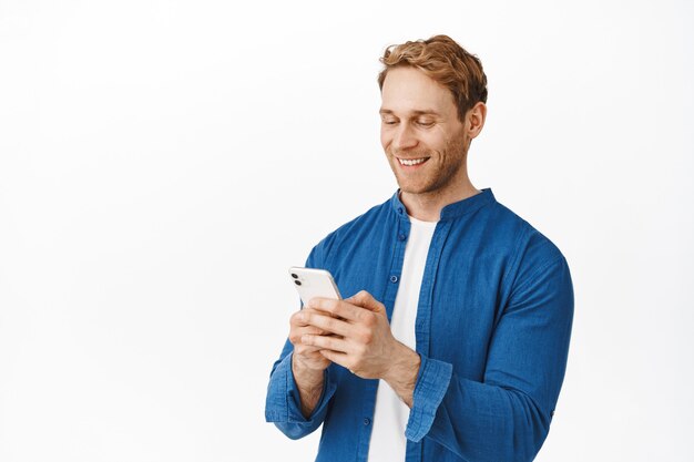 Moderner offener Kerl mit Telefon in den Händen, der chattet, Nachrichten liest oder den Bildschirm liest, während der Anwendung auf dem Smartphone-Display lächelt und über der weißen Wand steht