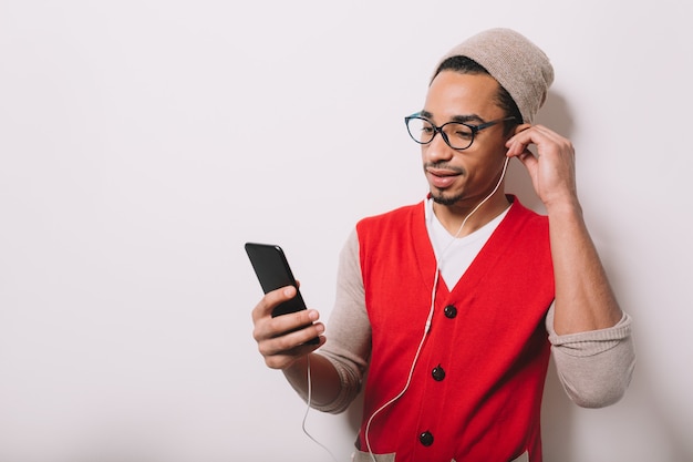 Moderner schöner afroamerikanischer Mann trägt Mütze und Brille hört Musik in Kopfhörern und hält Smartphone auf Grau