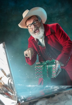 Moderner stilvoller weihnachtsmann in rotem modischem anzug und cowboyhut an dunkler wand