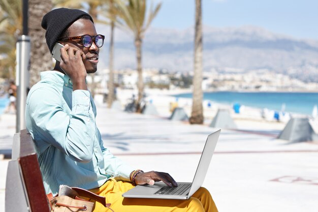 Moderner trendiger aussehender junger dunkelhäutiger Geschäftsmann, der entfernt auf Laptop-PC arbeitet und Handy für Geschäftsanrufe beim Sitzen auf Promenade entlang der blauen Küste am sonnigen Tag verwendet