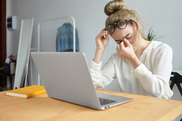 Modernes Technologie-, Job- und Personenkonzept. Porträt einer müden jungen Mitarbeiterin mit Haarknoten, die die Brille abnimmt und ihren Nasenrücken massiert und sich wegen viel Arbeit gestresst fühlt