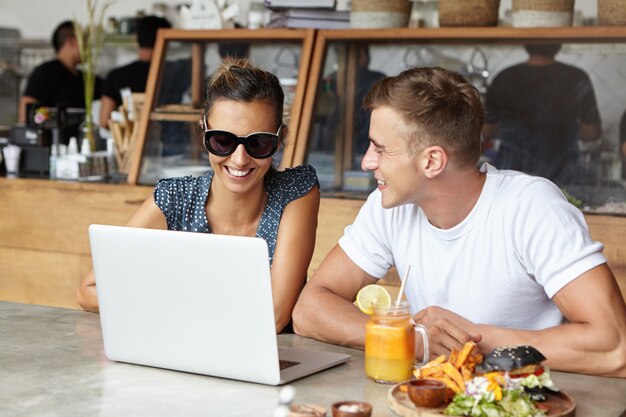 Modernes Technologie- und Kommunikationskonzept. Glückliches Paar, das Video anschaut oder Bilder im Internet mit kostenlosem Wi-Fi auf allgemeinem Laptop zusammen beim Mittagessen im Café durchsucht. Selektiver Fokus