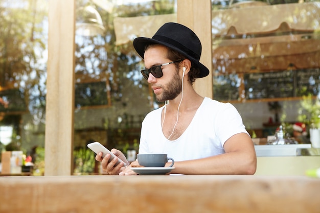 Modernes Technologie- und Kommunikationskonzept. Hübscher Student in den weißen Kopfhörern, die im Kaffeehaus im Freien entspannen Videoanruf machen