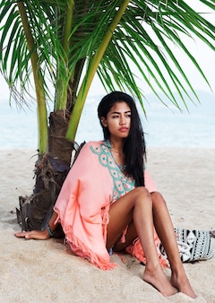Modesommerporträt des jungen schönen asiatischen modells, das am tropischen strand entspannt, boho-trendkleid trägt, auf weißem sand nahe palme sitzend.