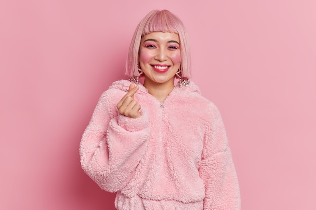 Modische asiatische Frau mit hellem Make-up macht Mini-Herzgeste koreanisch wie Zeichen lächelt angenehm hat rosa Haare und Pelzmantel posiert Innenkleider für Disco-Party. Körpersprachenkonzept.