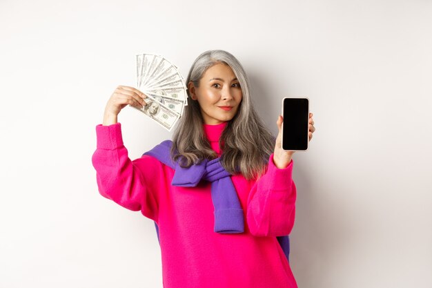 Modische asiatische Seniorin mit Gelddollar und leerem Smartphone-Bildschirm, Demonstration des Online-Shops, stehend auf weißem Hintergrund