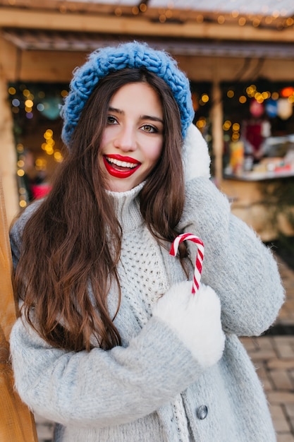 Modische brünette Frau in den weißen Handschuhen, die Winterzeit genießen. Außenporträt des raffinierten langhaarigen weiblichen Modells im stilvollen blauen Hut, der mit Vergnügen am kalten Morgen aufwirft, der Weihnachtssüßigkeit hält.