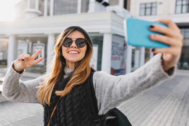 Modische charmante schöne Frau in der modernen Sonnenbrille, warmer Winterpullover, der Selfie-Porträt auf Straße im Stadtzentrum macht. Stilvoller Look, Spaß haben, positive, strahlende Emotionen ausdrücken.