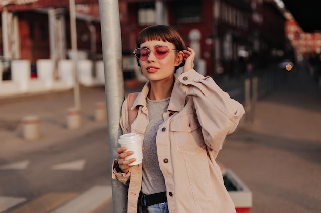 Modische Dame, die draußen eine Tasse Kaffee hält Brünettes Mädchen in beige Jeansjacke und rosa Brille posiert mit geschlossenen Augen in der Stadt