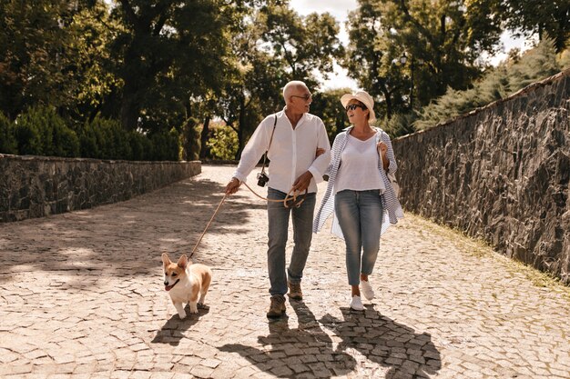 Modische Dame mit kurzen Haaren in gestreiftem Hemd, Jeans und Hut, die mit grauhaarigem Mann in heller Kleidung mit Hund im Park gehen.