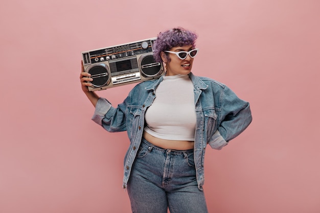 Modische Dame mit kurzen lila Haaren in weißer Sonnenbrille, in übergroßer Jeansjacke mit Retro-Radio.