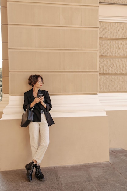 Modische Frau in weißer Hose, schwarzer Jacke und Stiefeln posiert im Freien Brunettemädchen mit Handtasche und Brille hält Telefon draußen
