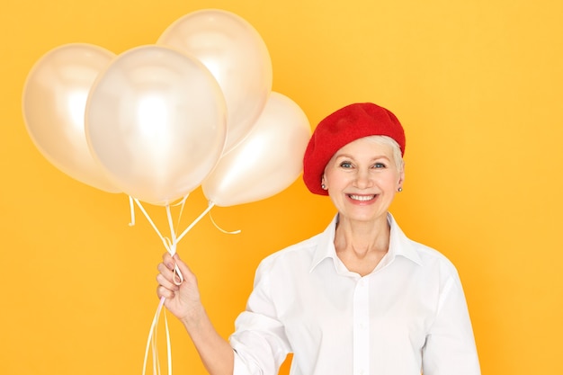 Modische fröhliche europäische Rentnerin in weißem Hemd und roter Haube, die Heliumballons hält und lächelt, Jubiläum oder Geburtstag feiert, glücklichen überglücklichen Gesichtsausdruck hat