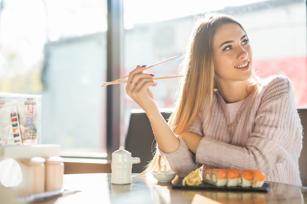 Modische junge blonde Frau im weißen Pullover, der Sushi zum Mittagessen an einem kleinen Kaffee isst