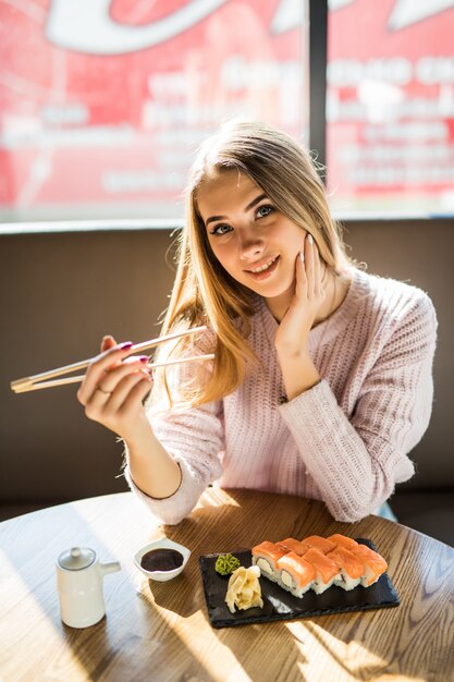 Modische junge blonde Frau im weißen Pullover, der Sushi zum Mittagessen an einem kleinen Kaffee isst