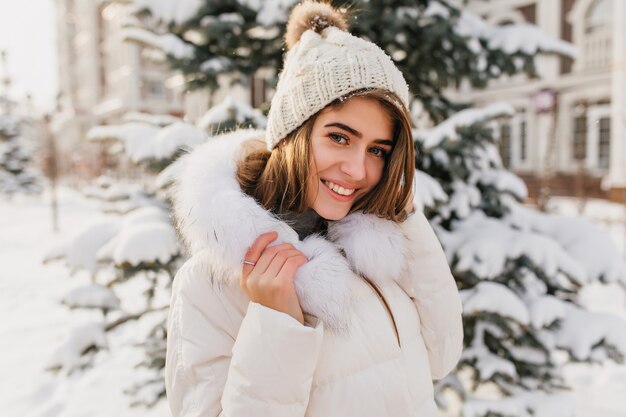 Modische junge Frau in der weißen Strickmütze, die freundlich auf der Straße voll mit Schnee lächelt. Erstaunliche europäische Frau, die Winterzeit genießt