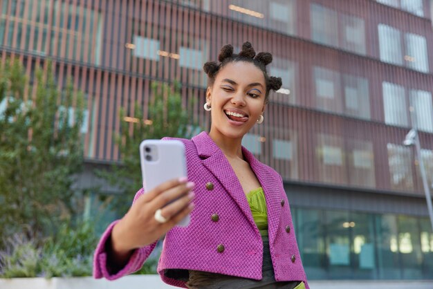 Modische junge Frau mit Brötchenfrisur zwinkert Augenposen für Selfie an der Smartphone-Kamera trägt modische rosafarbene Jackenposen im Freien in urbaner Umgebung hat fröhlichen Ausdruck Street Lifestyle