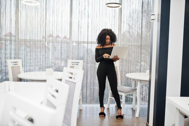 Modische junge schöne afrikanisch-amerikanische Geschäftsfrau mit Afro-Frisur trägt einen eleganten schwarzen Ständer mit Laptop zur Hand