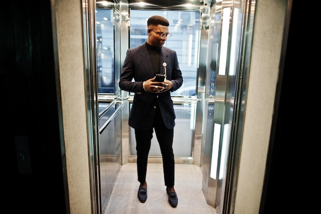 Modischer afrikanisch-amerikanischer Mann in Anzug und Brille mit Handy und Tasse Kaffee in der Hand posiert im Aufzug