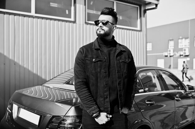 Modischer arabischer Mann trägt schwarze Jeansjacke und Sonnenbrille, die sich gegen ein modernes Geschäftsauto stellen Stilvoller, erfolgreicher und modischer arabischer Modelltyp